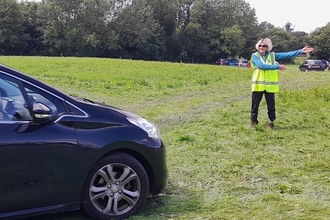 Marshal volunteer directing cars to a parking space in a field