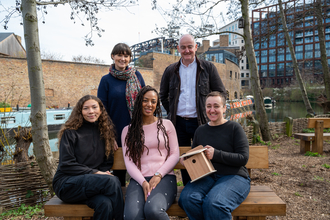 A woman and a man stood behind three women sat on a bench. 