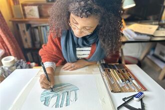 Jamey is sat at a table, adding detail to a watercolour on a piece of paper with a fine liner