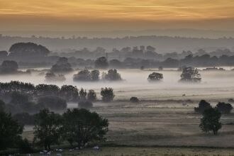 The flat landscape of the Somerset Levels at sunrise, as seen from Burrow Mump. Trees and hedgerows are silhouetted against the pale light, with mist in the midground and a dark orange sky in the background.