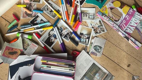messy scattered pens, polaroid photos, coloured card and people's feet in shoes on wooden floorboards