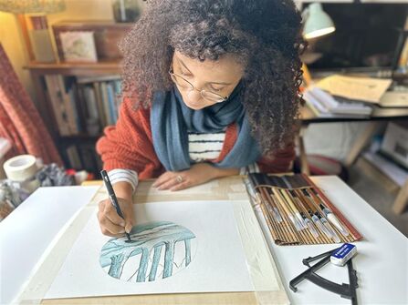 Jamey is sat at a table, adding detail to a watercolour on a piece of paper with a fine liner